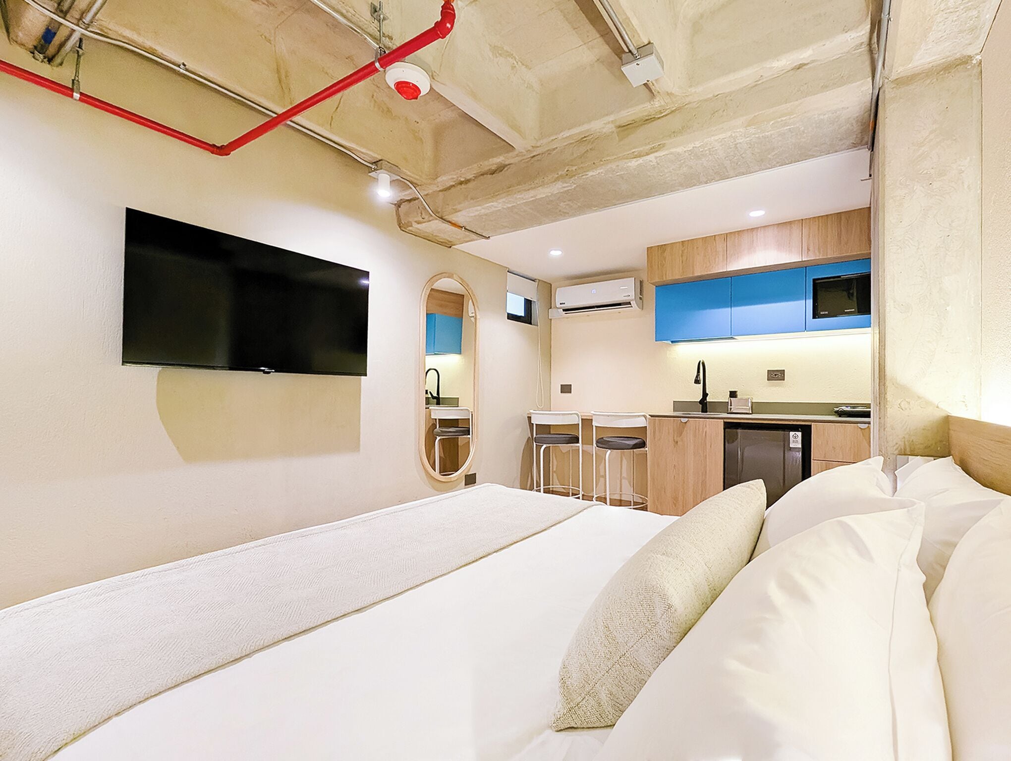 A compact hotel room with a bed, a TV mounted on the wall, a small kitchenette, and a bathroom with a mirror and sink.
