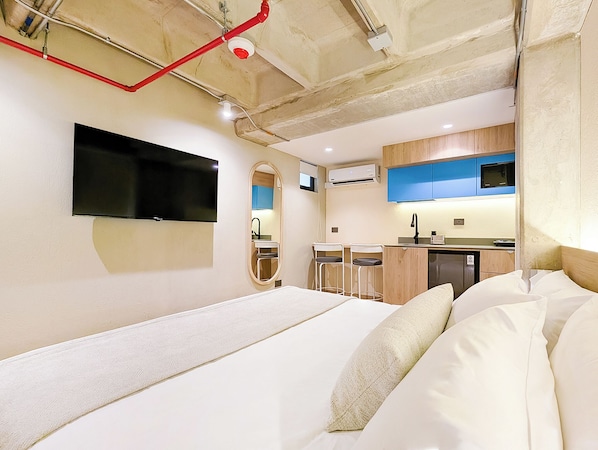 Habitación de hotel compacta con cama, televisión montada en la pared, pequeña kitchenette y baño con espejo y lavamanos.