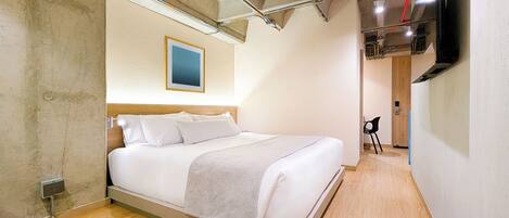 A modern hotel room with a bed, a mounted TV, and exposed ceiling pipes.