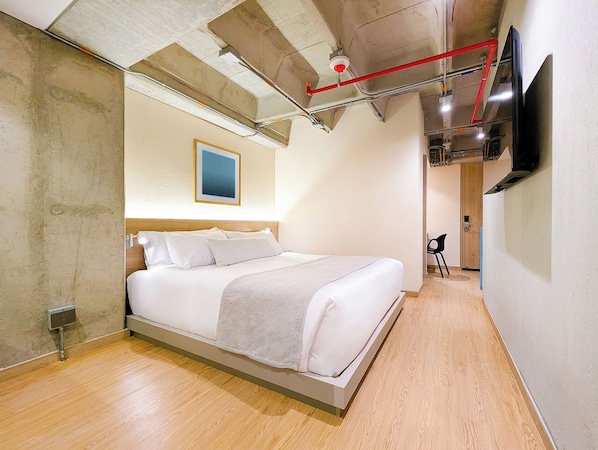 Una habitación de hotel moderna con cama, televisor montado y tuberías del techo a la vista.