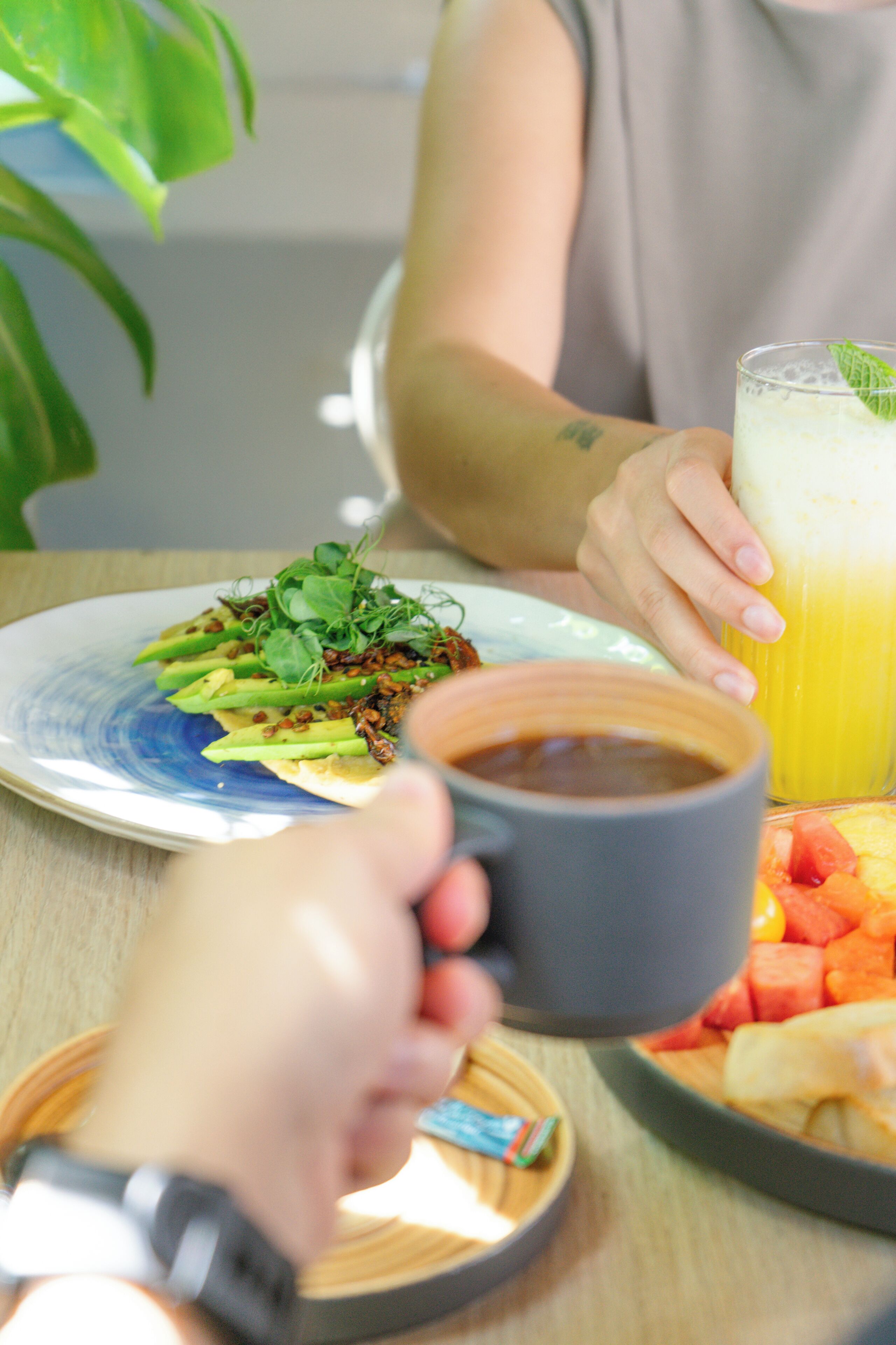 A person holding a mug with a beverage, a plate with food, and a glass of juice on a table.