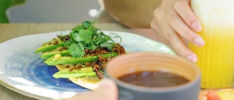 A person holding a mug with a beverage, a plate with food, and a glass of juice on a table.