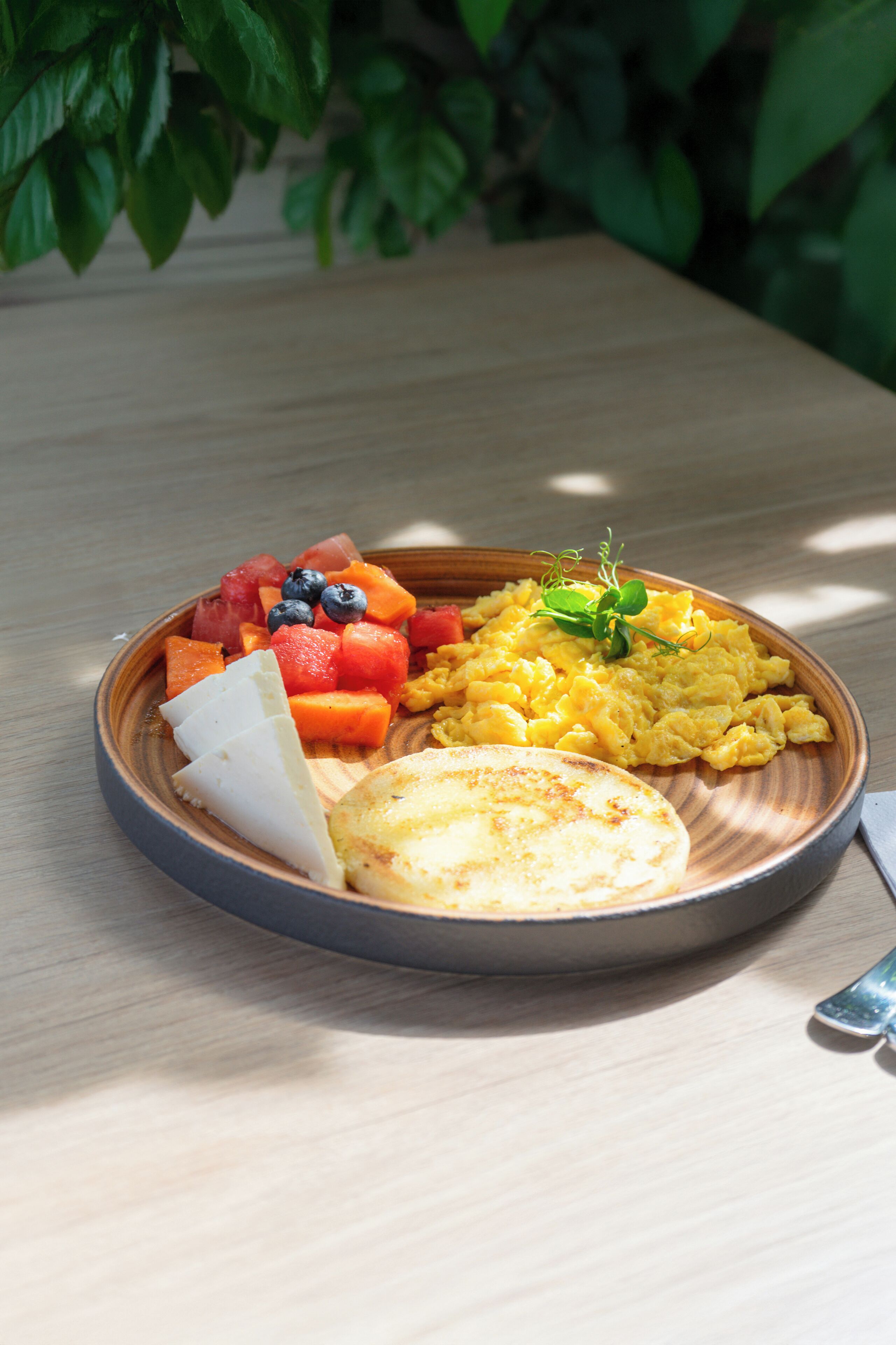 A wooden table with a plate of food including scrambled eggs, fruit, and a piece of bread.