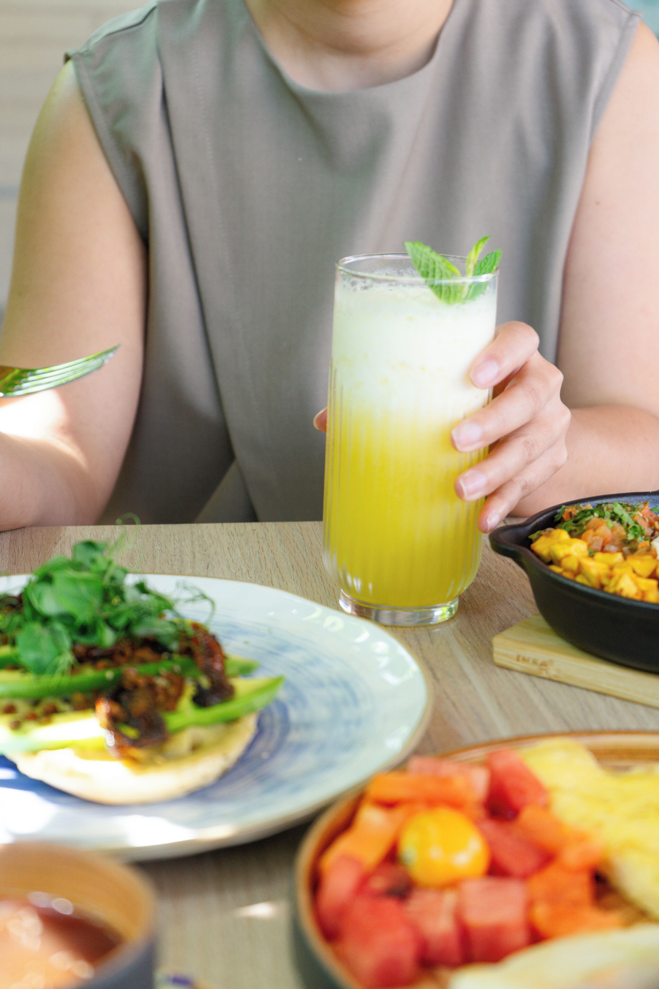 Una persona sosteniendo un vaso alto de una bebida amarilla, un plato con ensalada y un tazón de fruta.