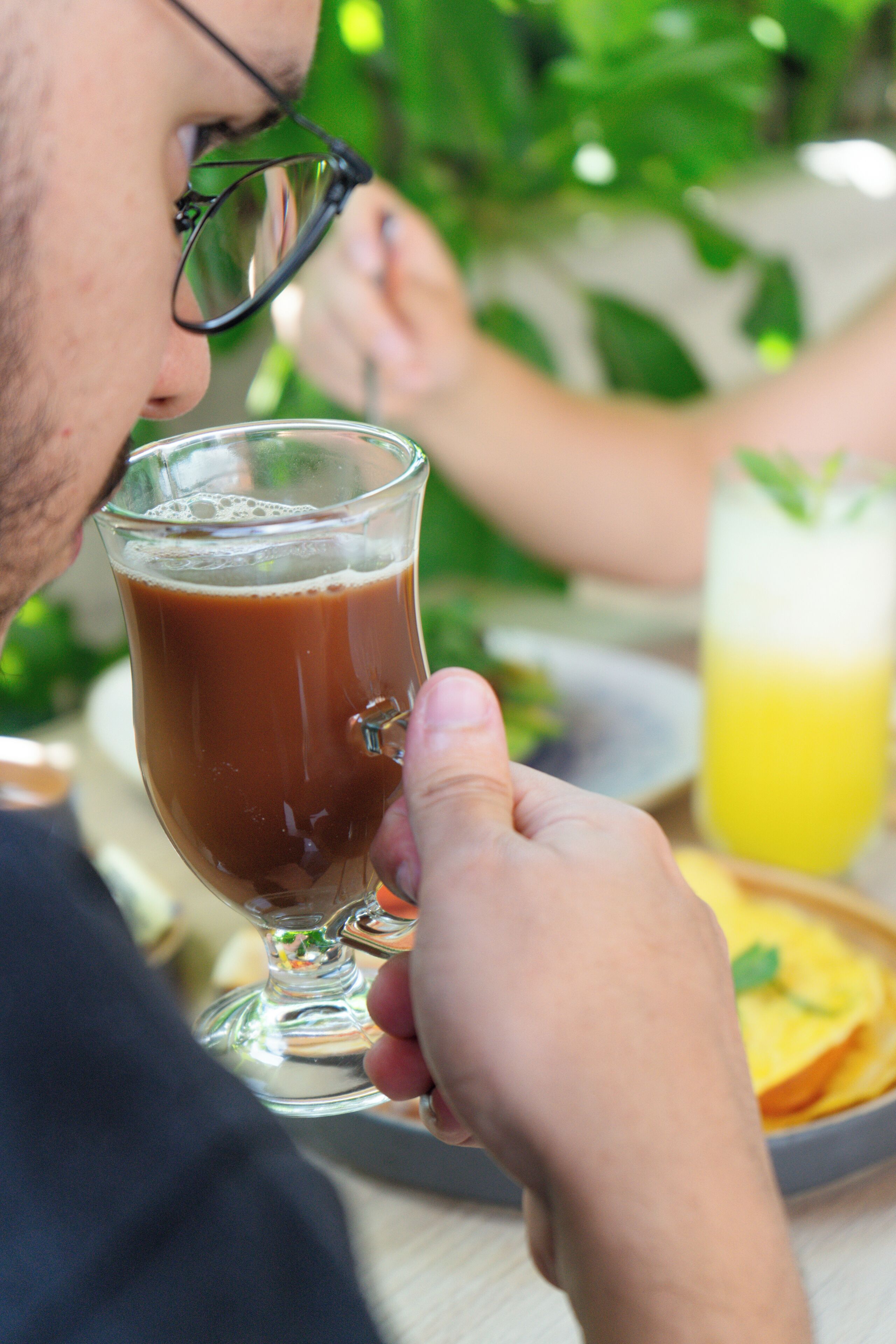 A person holding a glass of coffee, with a plate of food and a glass of juice in the background.