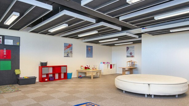 A playroom with a carpeted floor, a round white playpen, a red storage unit, and a pegboard with children's items.