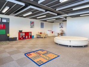 Una sala de juegos con piso alfombrado, un corralito blanco redondo, un mueble de almacenaje rojo y un tablero de anuncios con objetos infantiles.