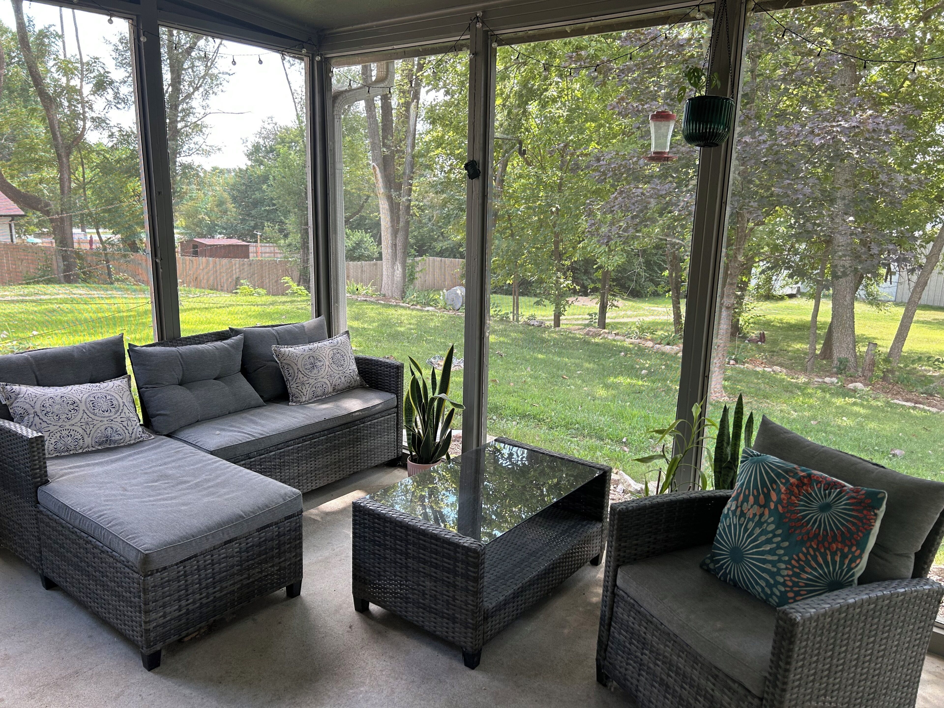 A covered patio with wicker furniture, a glass coffee table, and a view of a backyard with grass and trees.