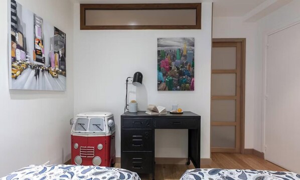 Une pièce comprenant un lit, un bureau avec une lampe, un vieux réfrigérateur rouge et deux tableaux accrochés au mur.