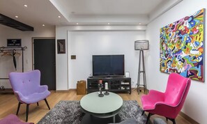 Un salon moderne avec un téléviseur à écran plat, deux fauteuils, une table basse et un tableau abstrait aux couleurs vives accroché au mur.