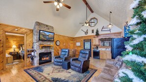A cozy log cabin interior with a fireplace, leather recliners, a large rug, and a Christmas tree.
