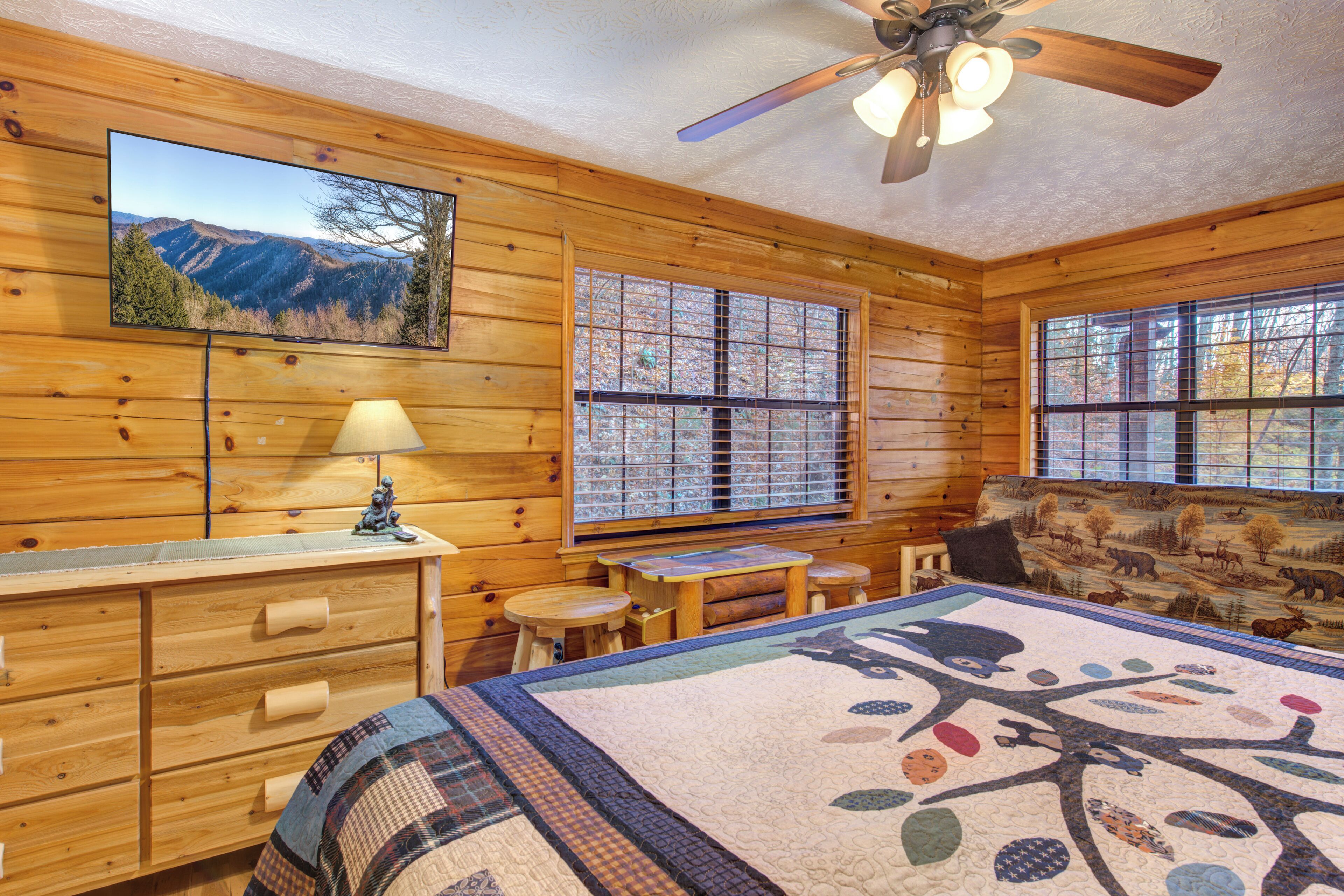 Une chambre de style rustique, aménagée dans une cabane en rondins, comprenant un lit, un bureau, une chaise, une fenêtre donnant sur les montagnes et un ventilateur de plafond.