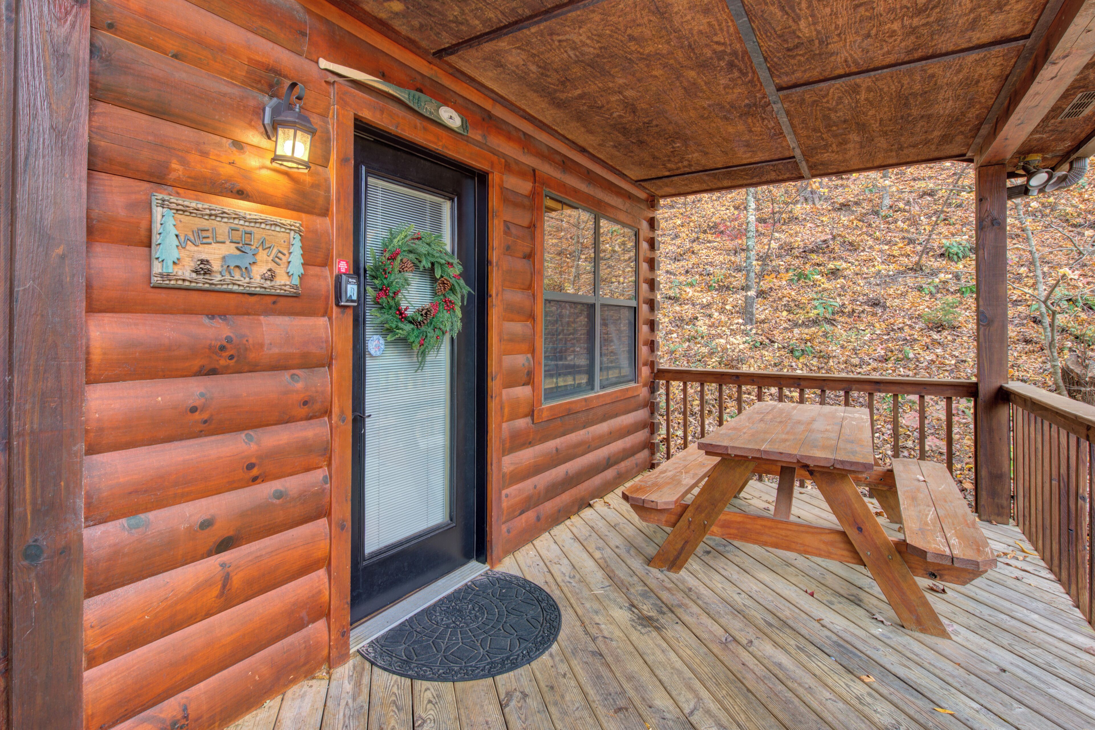 A wooden cabin with a front door, a welcome sign, a wreath, and a picnic table on the porch.