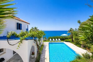 Uma piscina com vista para o mar, um edifício branco com detalhes azuis e uma fileira de espreguiçadeiras brancas.