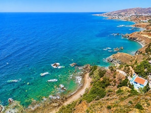 A coastal landscape with clear blue waters, rocky outcrops, and a path leading to a building with a red roof.