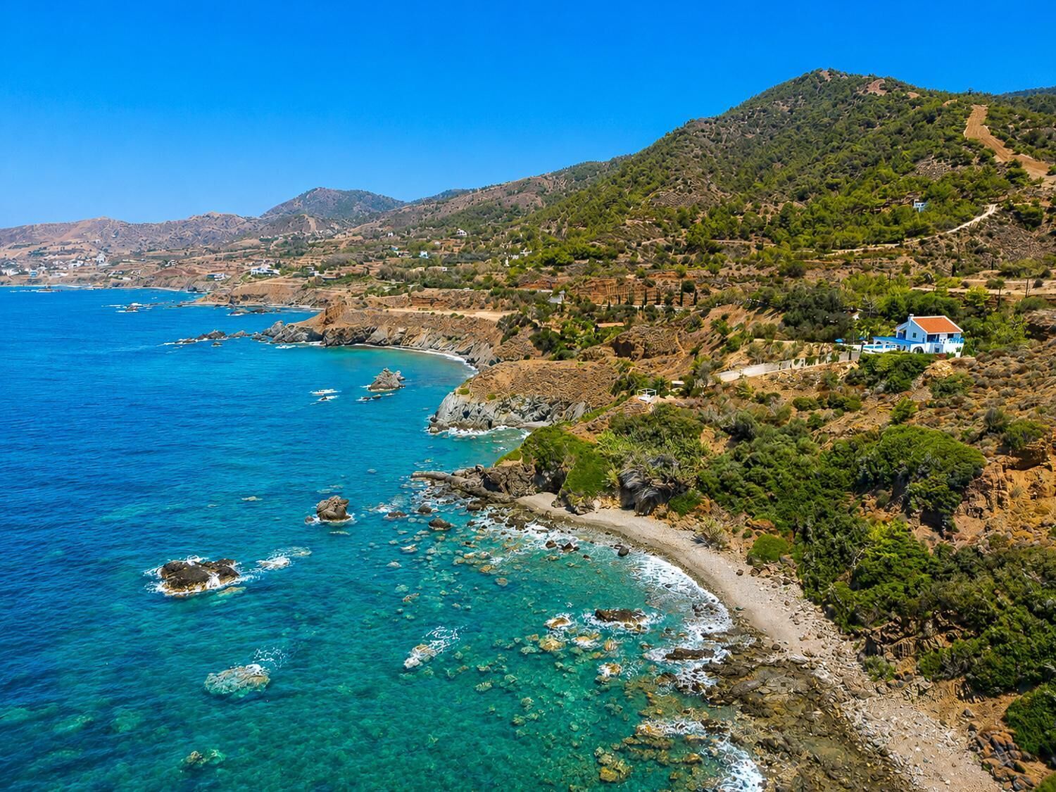 A coastal landscape with clear blue waters, rocky shores, and a hillside with sparse vegetation and a few buildings.