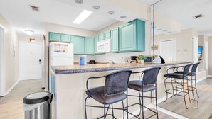 A modern kitchen with teal cabinets, a central island with bar stools, and a stainless steel trash can.