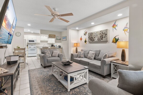 A modern living room with a grey sofa set, a coffee table, and a TV. There is a kitchen in the background with white cabinets and a microwave.