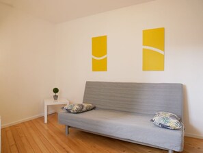 Una habitación moderna con un sofá gris, dos almohadas, una mesita de noche blanca con una planta y dos cuadros amarillos en la pared.