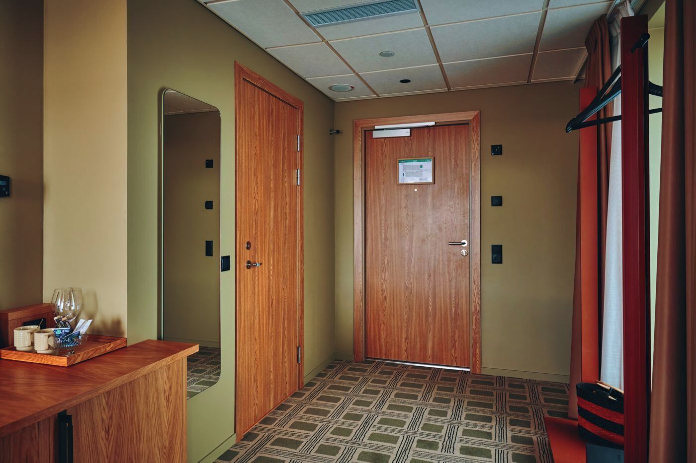 A hotel hallway with two wooden doors, a reception desk with a glass container, and patterned carpet.