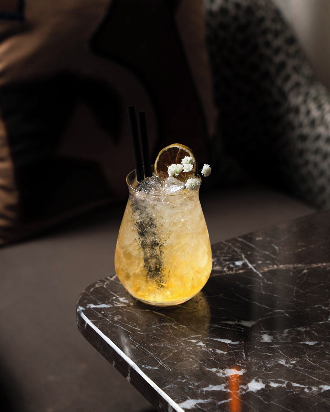 A cocktail in a glass with ice, garnished with a slice of orange and white flowers, on a marble surface.