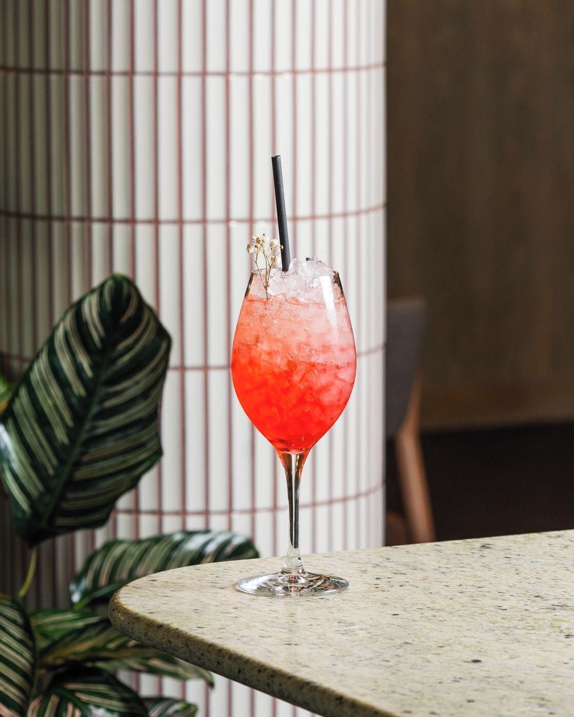 A cocktail in a glass with a straw, ice cubes, and a decorative flower on top, placed on a table with a plant in the background.