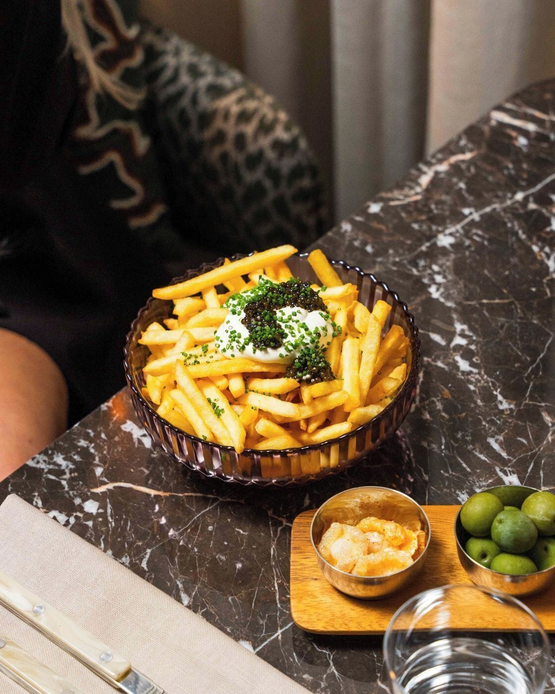 A bowl of French fries topped with sour cream and caviar, accompanied by a small bowl of cheese and a glass of water on a marble table.
