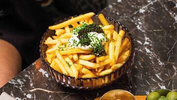 A bowl of French fries topped with sour cream and caviar, accompanied by a small bowl of cheese and a glass of water on a marble table.