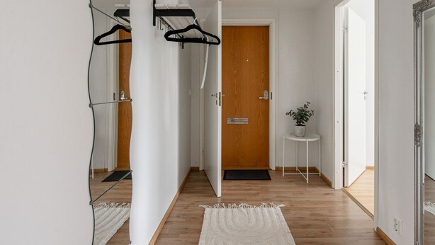 Un pasillo con suelo de madera, una puerta blanca, una pequeña mesa blanca con una planta y un espejo que refleja el pasillo.