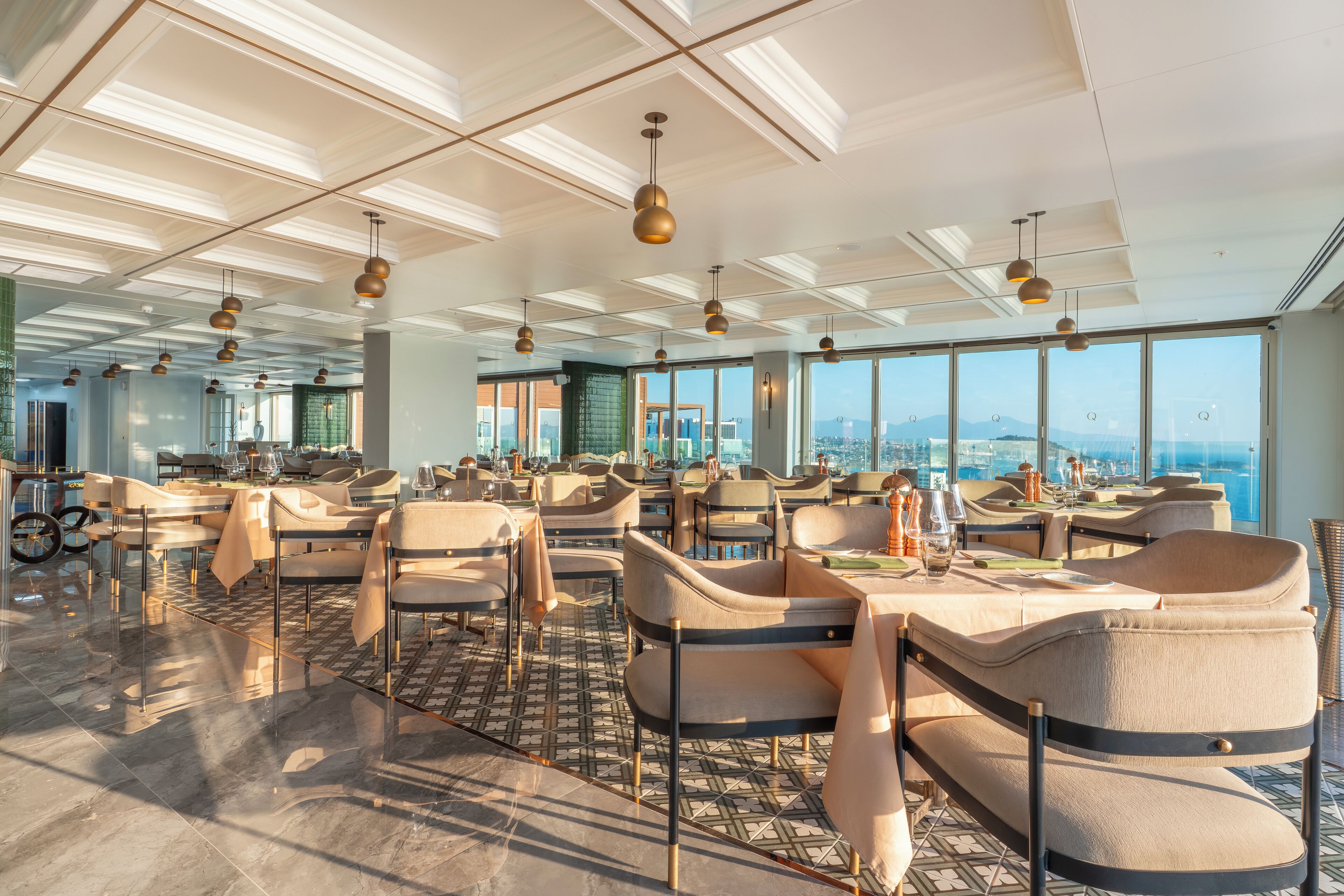 Un restaurant spacieux avec de hauts plafonds, un éclairage moderne suspendu et de grandes fenêtres offrant une vue sur la ville.