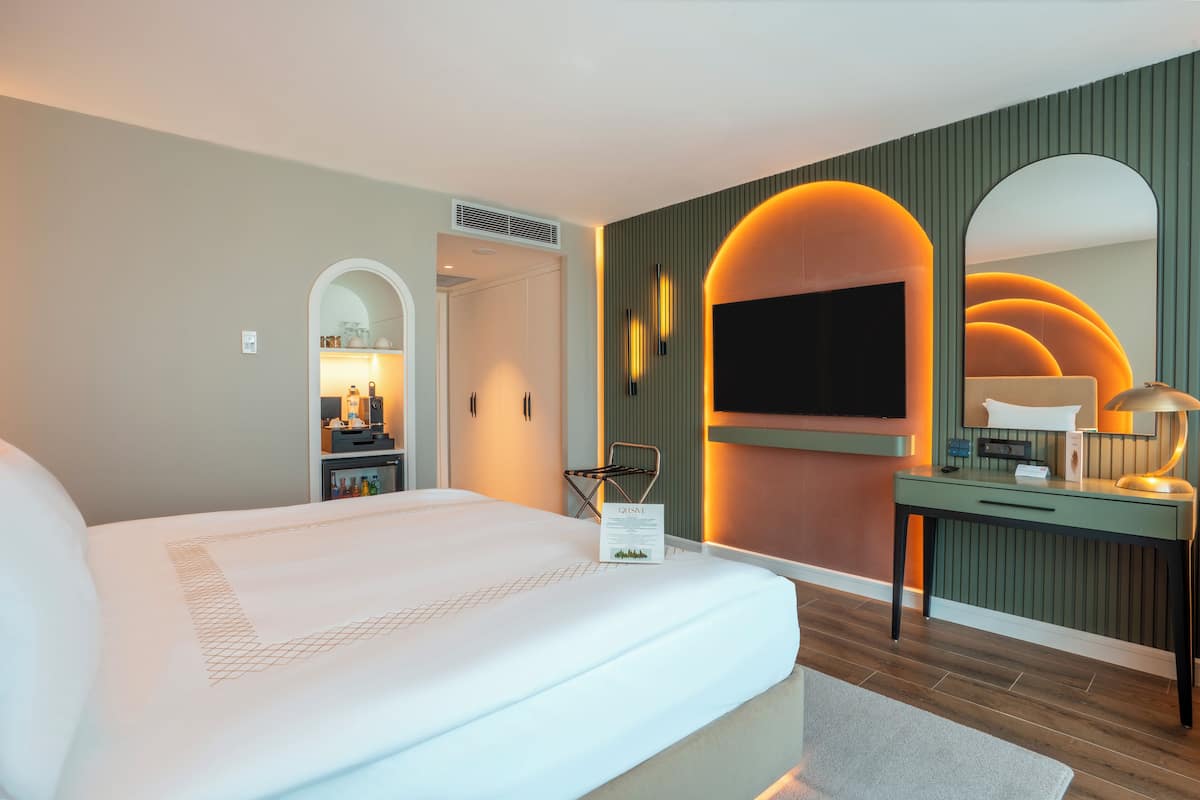 A modern hotel room with a large bed, a desk with a computer, and a TV mounted on the wall.