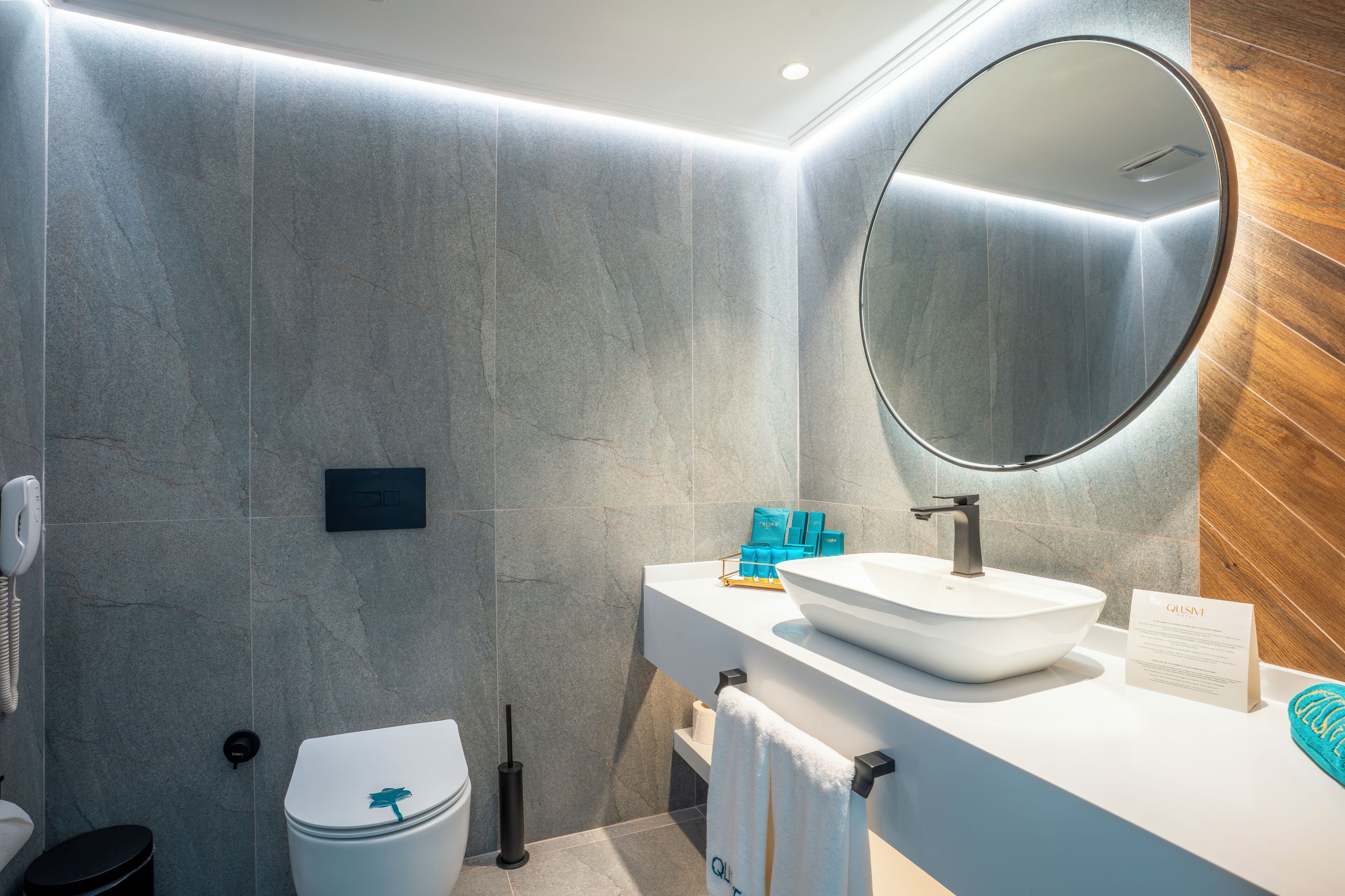 A modern bathroom with a white sink, a round mirror, and a wooden accent wall.