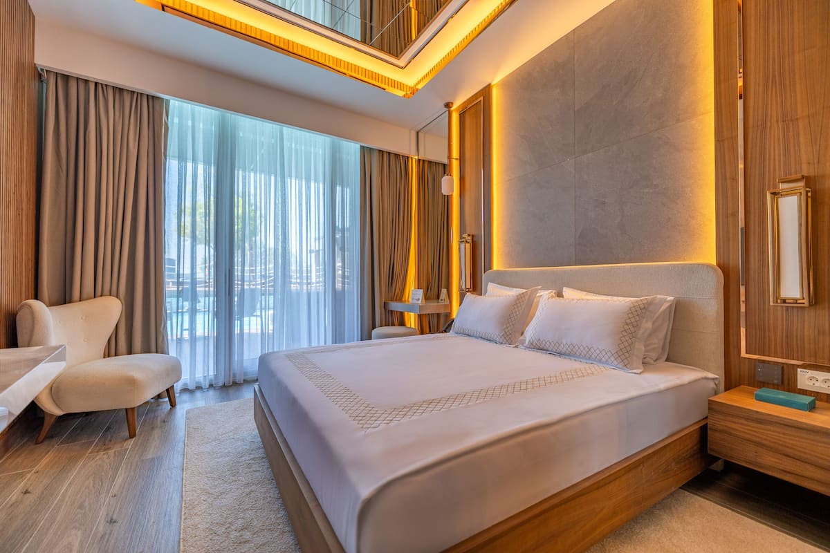 A modern hotel room with a large bed, a seating area with a chair, a bedside table, and a large window with curtains.
