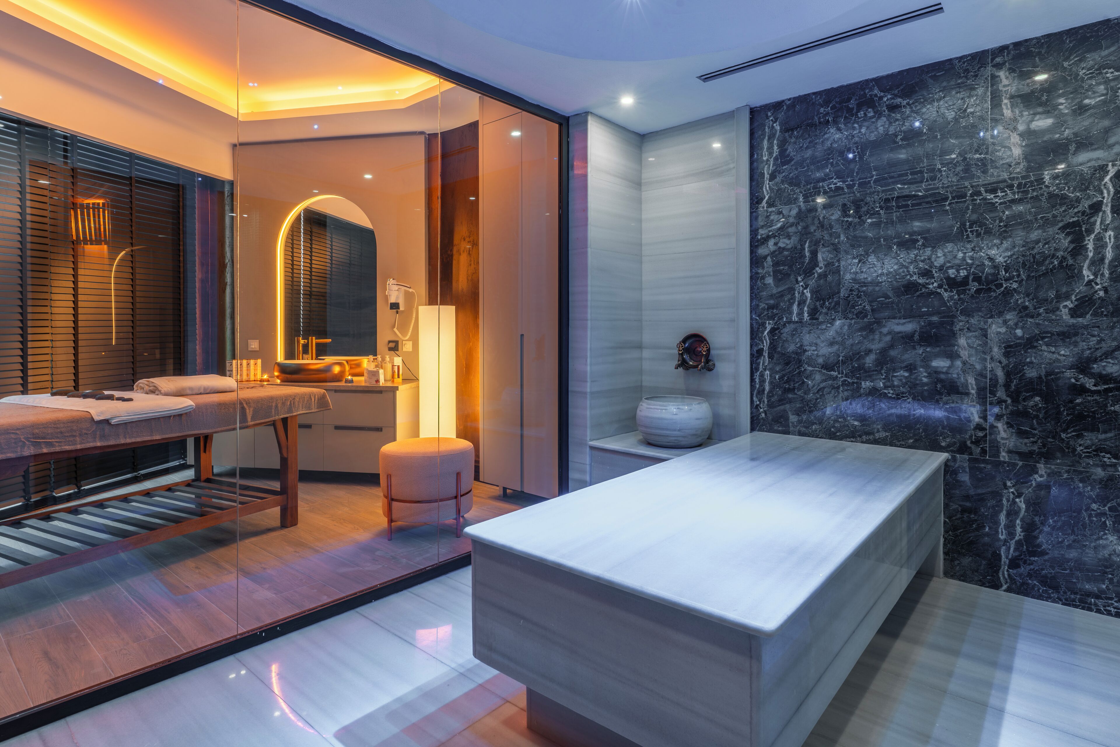 A modern bathroom with a large mirror, a wooden bench, a marble countertop, and a black marble wall.