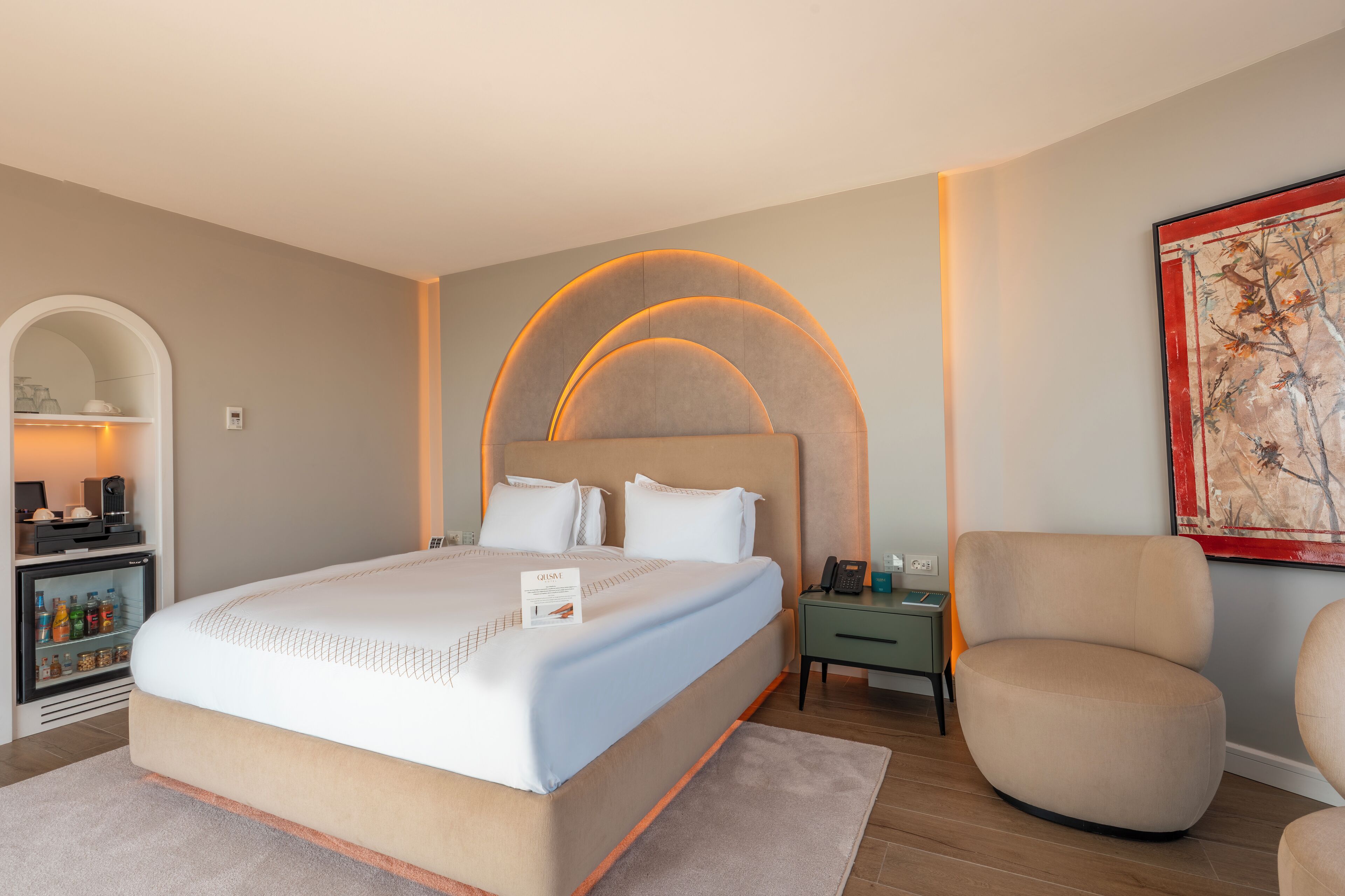 Una habitación de hotel moderna con una cama grande, una mesita de noche con lámpara, una silla y un cuadro en la pared.