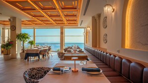 A restaurant with a long bench seating area, tables with chairs, and a view of the sea. - Qlusive Hotel (Kusadasi)