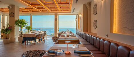Un restaurante con una larga zona de asientos tipo banco, mesas con sillas y vistas al mar.