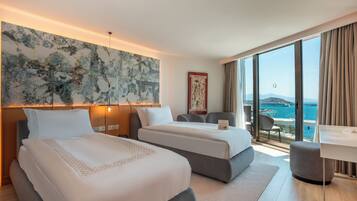 Habitación de hotel con dos camas, un escritorio y un amplio ventanal con vista al mar.