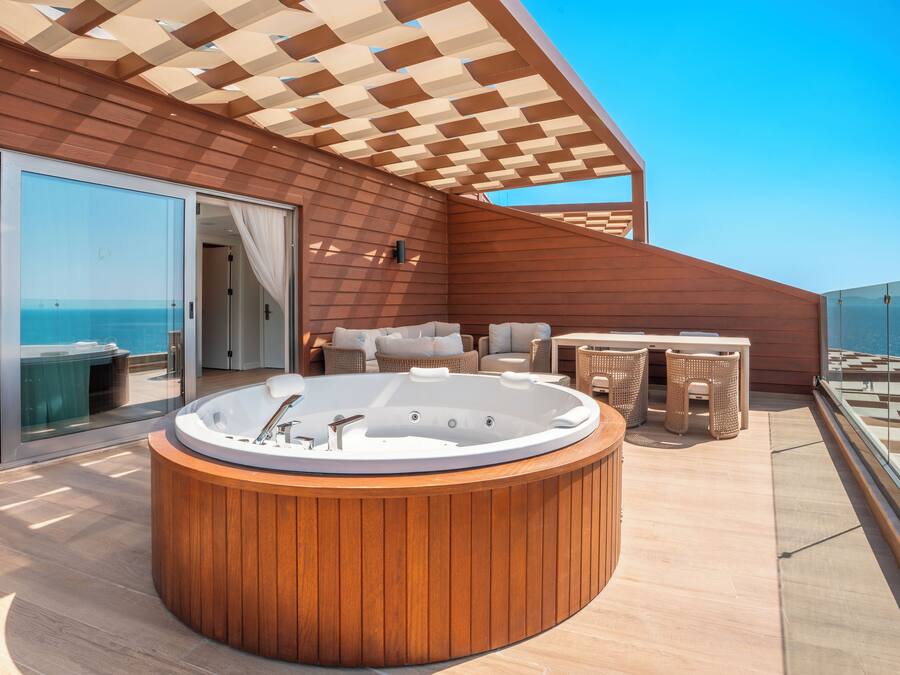 Una terraza de madera con un jacuzzi, mobiliario de mimbre y un ventanal que da a un balcón con vistas al océano.
