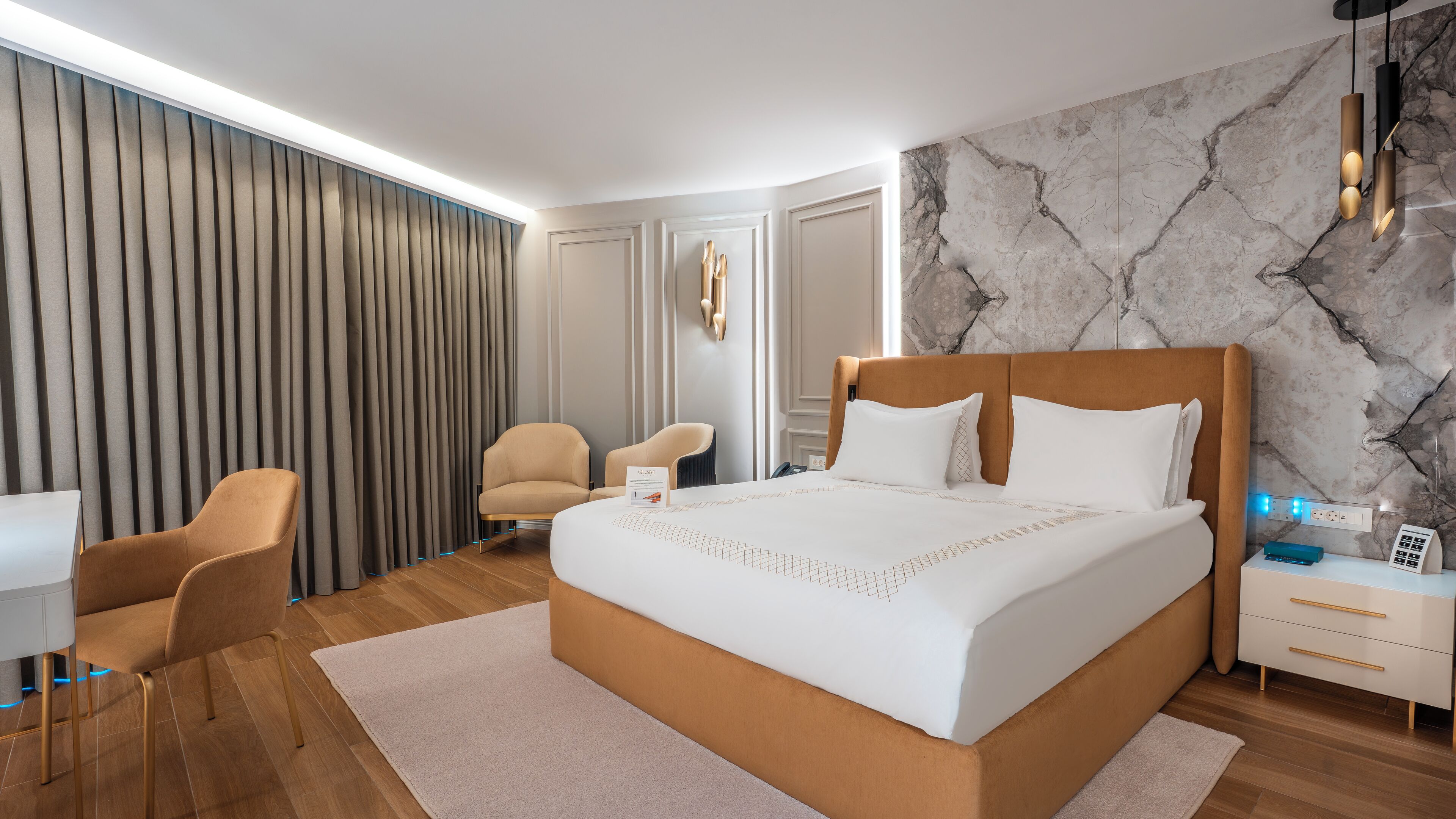 A modern hotel room with a large bed, two armchairs, a desk, and a marble-patterned wall.