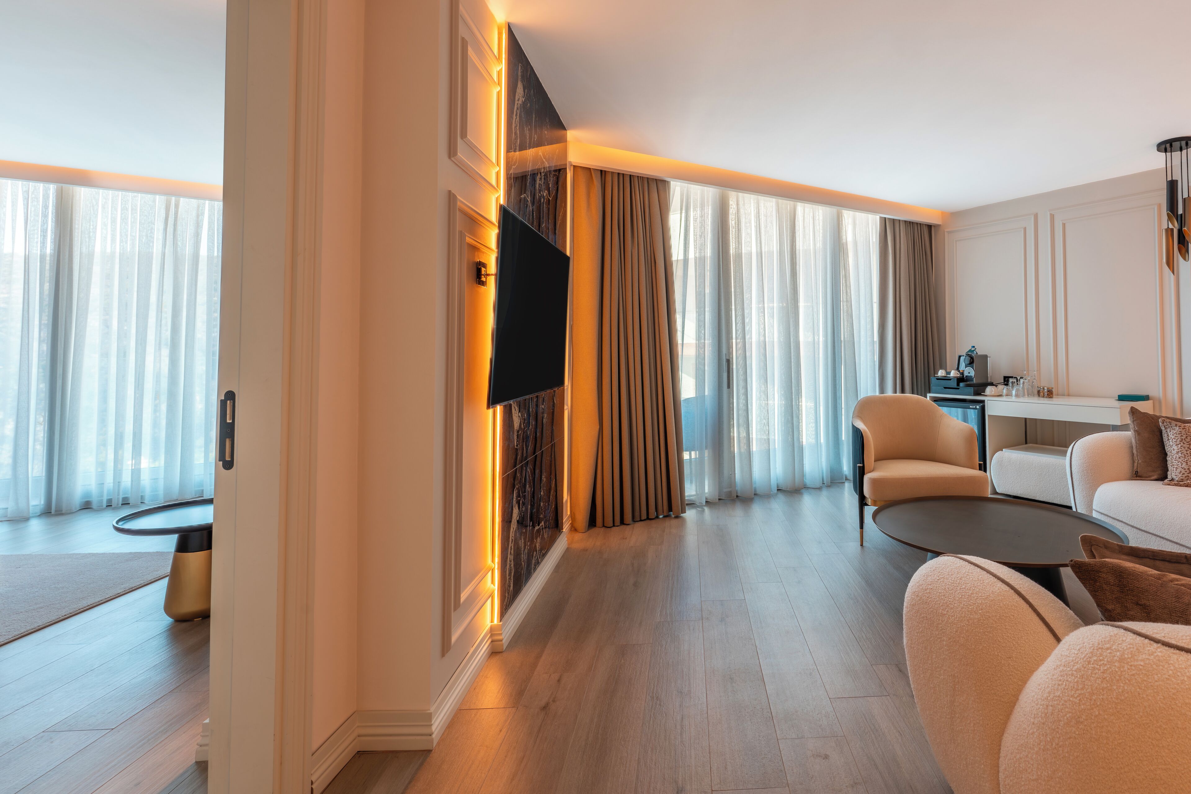 Quarto de hotel moderno com televisão de tela plana, sofá, mesa de centro e área de refeições com mesa e cadeiras.