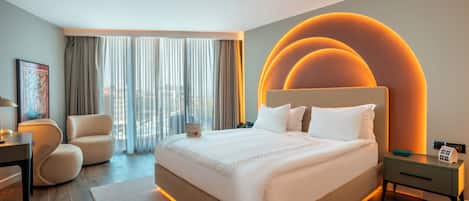 A modern hotel room with a large bed, a desk, a chair, and a view of the city through large windows.