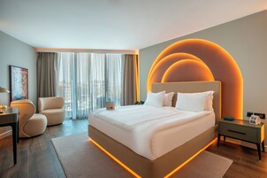 Quarto de hotel moderno com uma cama grande, uma escrivaninha, uma cadeira e vista para a cidade através de amplas janelas.
