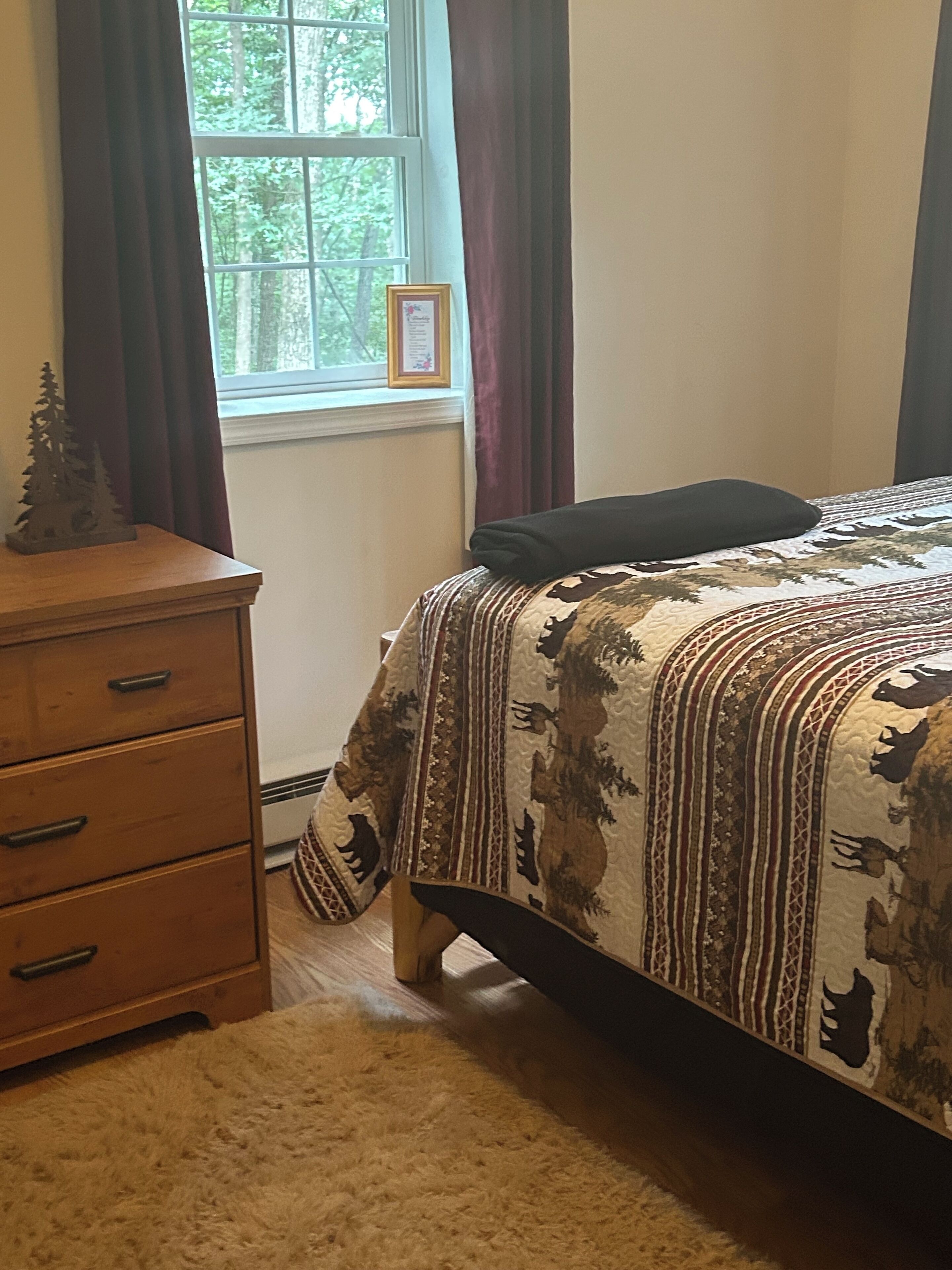 Ein Schlafzimmer mit Bett, Kommode, Fenster mit Vorhängen und einem gerahmten Bild an der Wand.