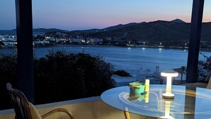 Un espace extérieur aménagé avec des chaises en osier et une table en verre, offrant une vue sur une étendue d’eau et des collines lointaines.