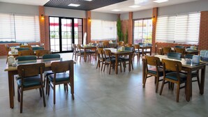 A spacious dining area with multiple tables and chairs, large windows with blinds, and a brick accent wall. - Azak Blue Otel (Alanya)