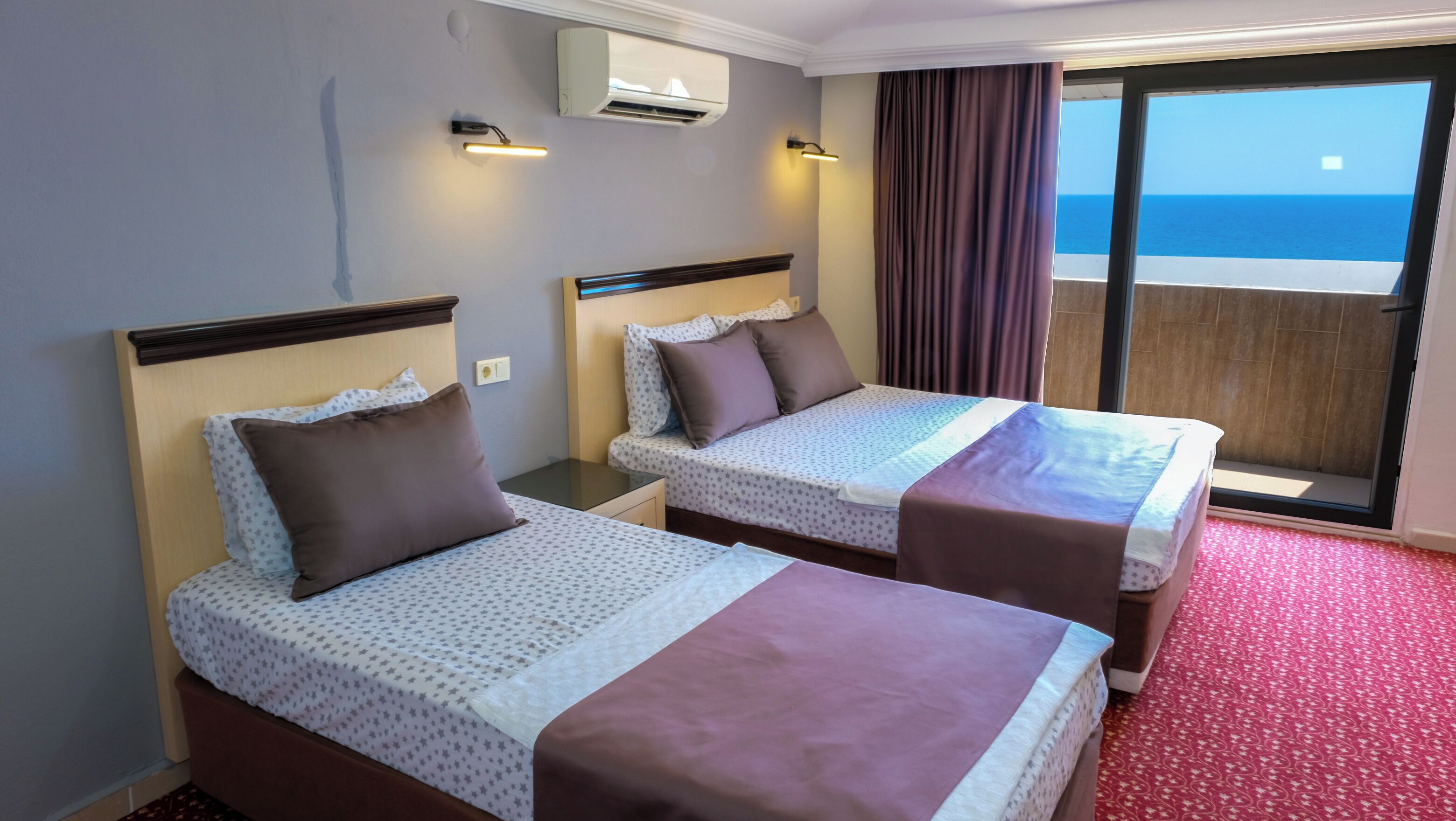 A hotel room with two beds, a balcony with a view of the sea, and an air conditioning unit on the wall.