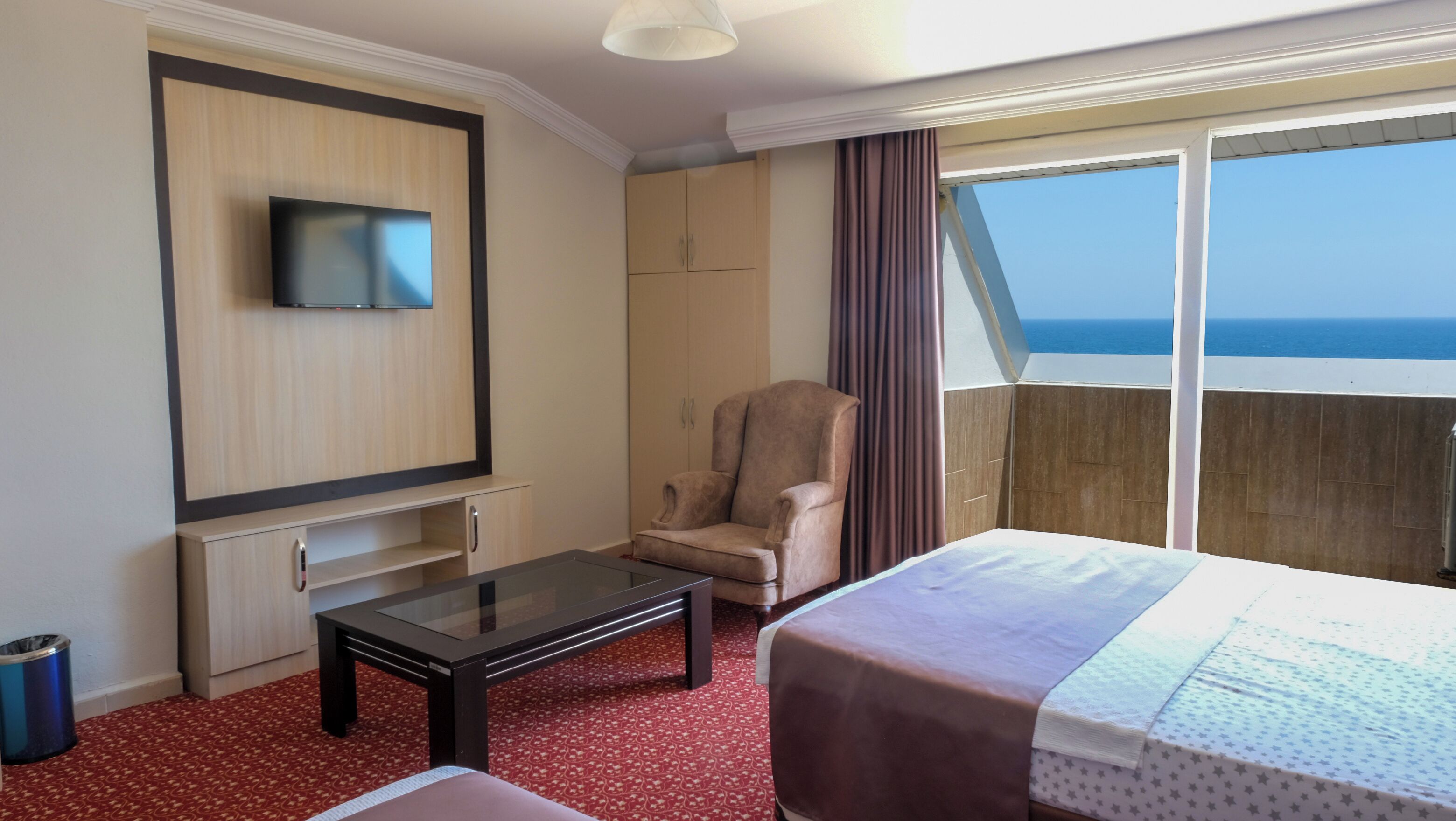 A hotel room with a bed, a TV mounted on the wall, a chair, a small table, and a view of the ocean.