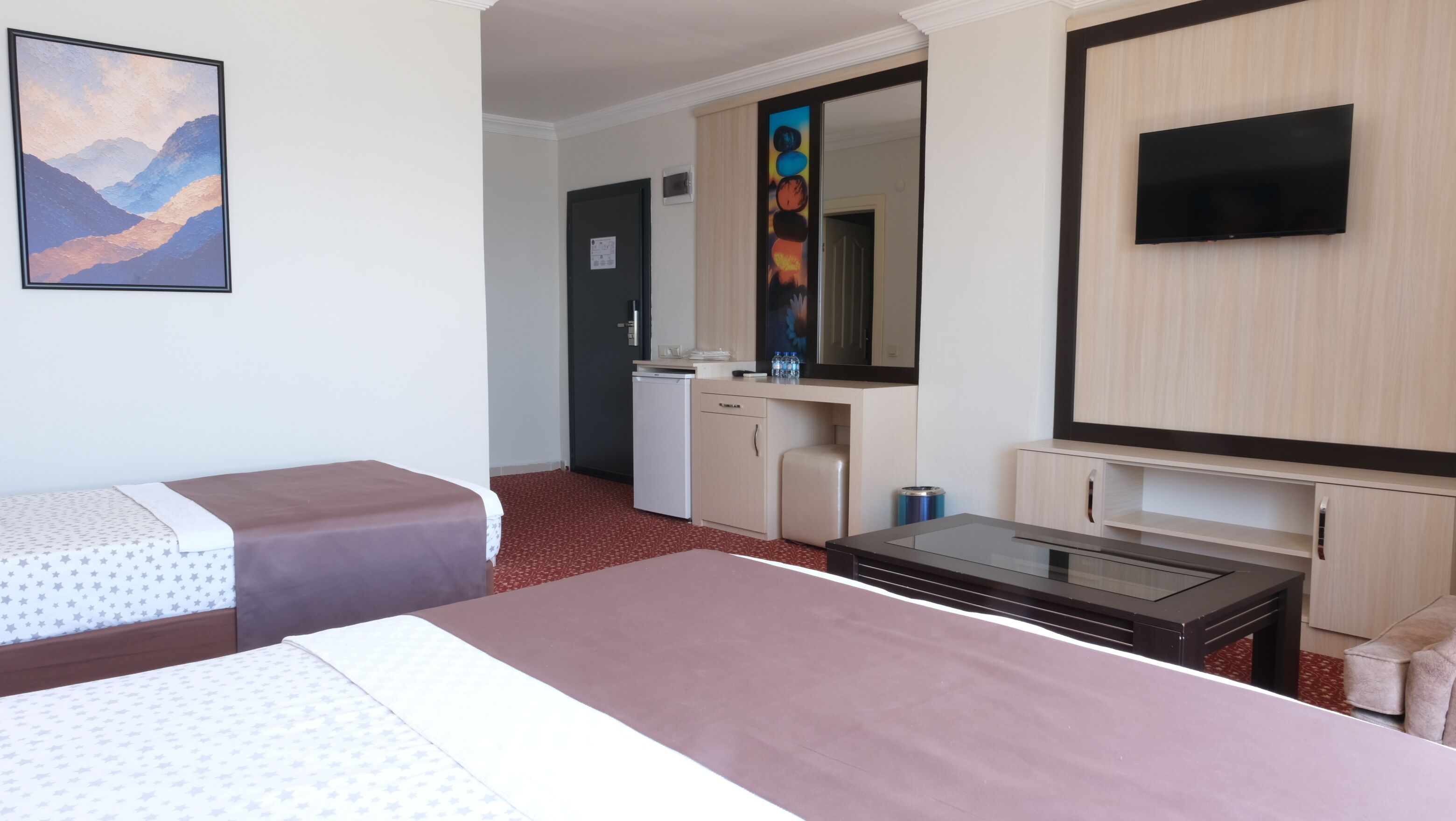 A hotel room with two beds, a desk, a TV, a minibar, and a bathroom visible in the background.