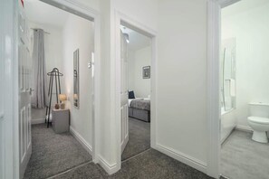 A hallway with a bed, a bedside table, and a lamp. There is a bathroom with a toilet and a mirror. - Ruby House - 2 Bedroom Huddersfield Yorkshire Flat (West Yorkshire)